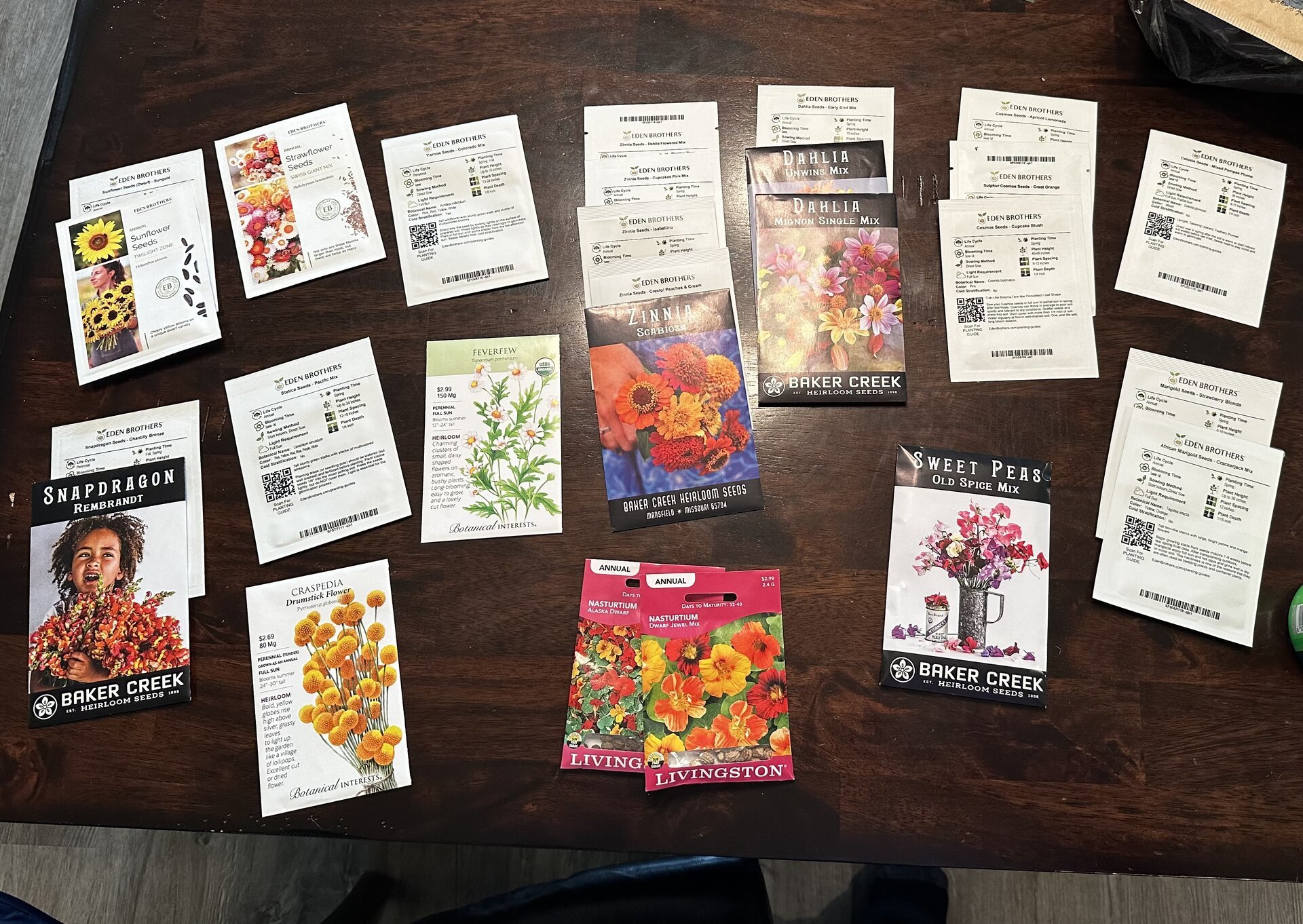Colorful 2026 seed collection spread on table showing various seed packets from Eden Brothers, Baker Creek, Botanical Interests, and Livingston seed companies